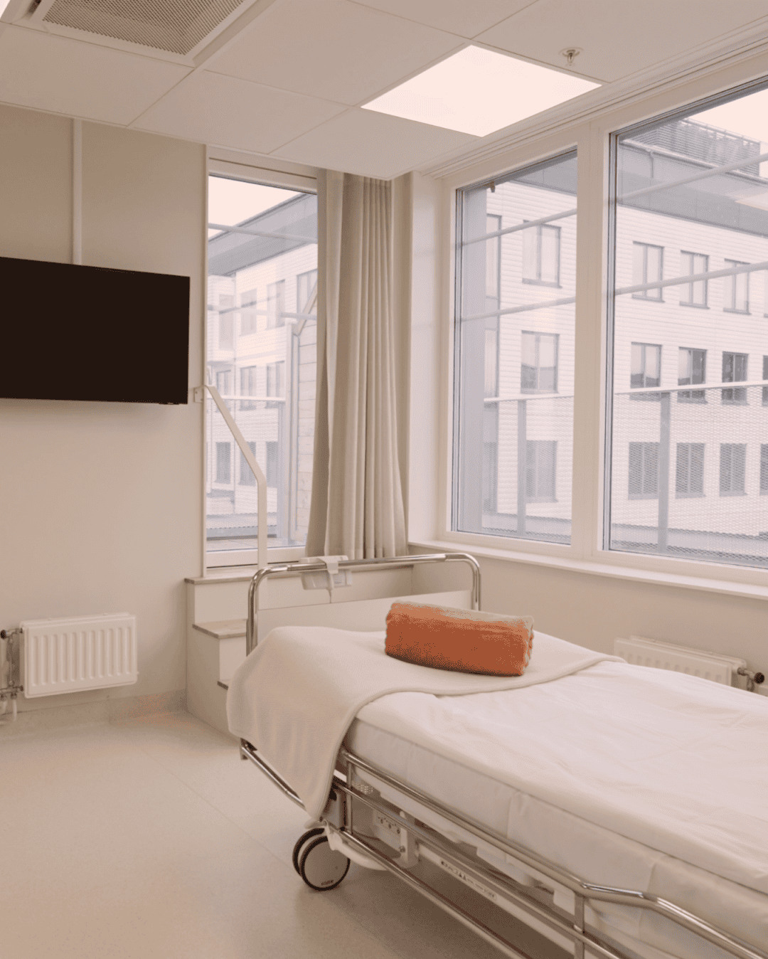 Säng i ett tomt sjukhusrum med stora fönster och TV på väggen.