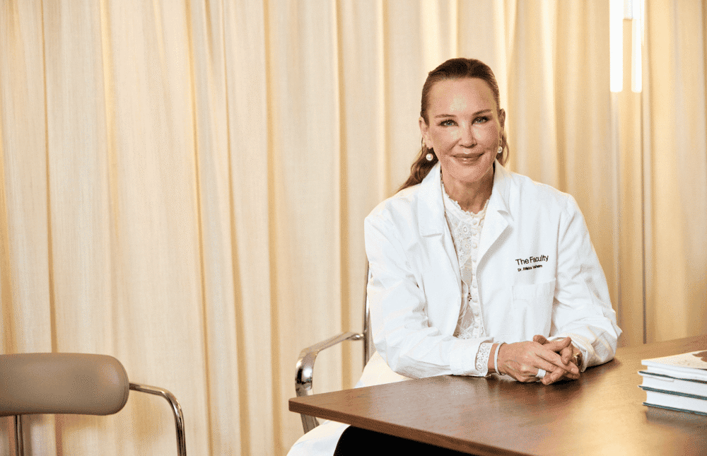 Kvinna i vit labbrock med The Faculty-logga, leende mot kameran.
